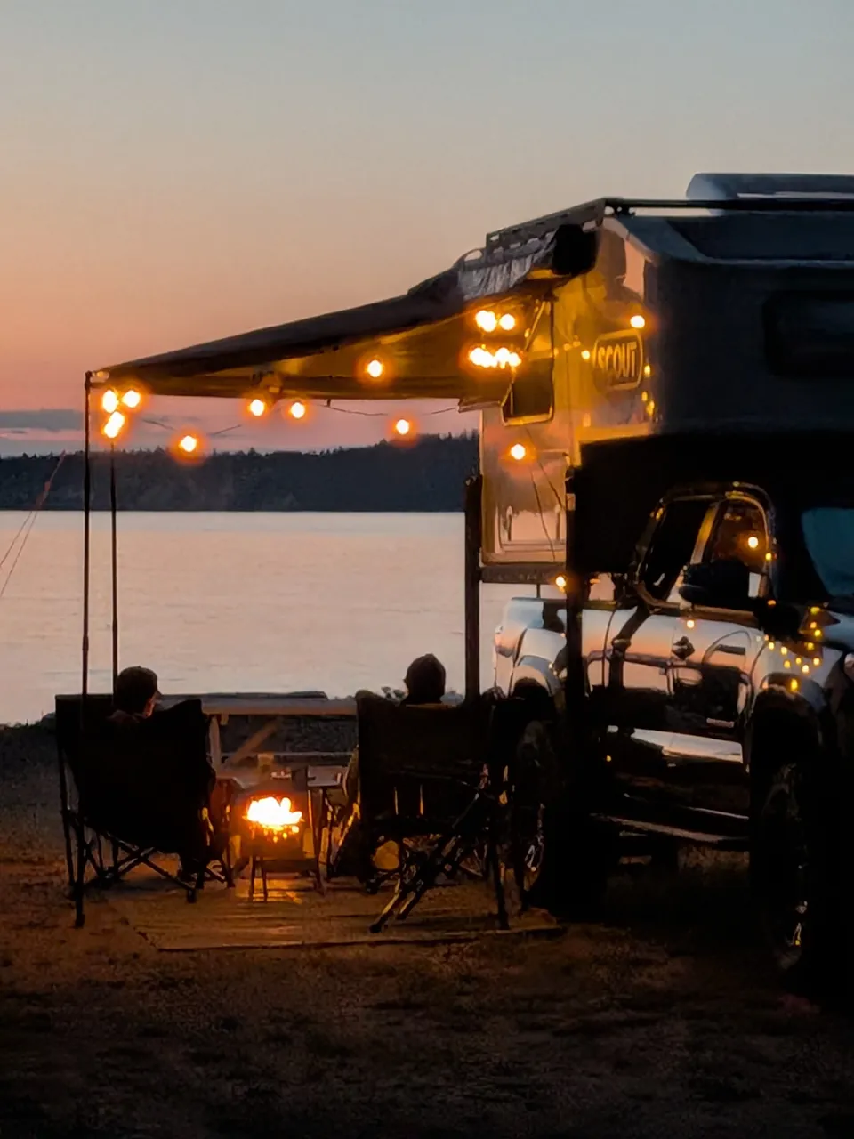 Twee mensen ontspannen naast een pick-upcamper met warme lichtjes, met uitzicht op het water in de schemering, bij Beachcomber RV Park