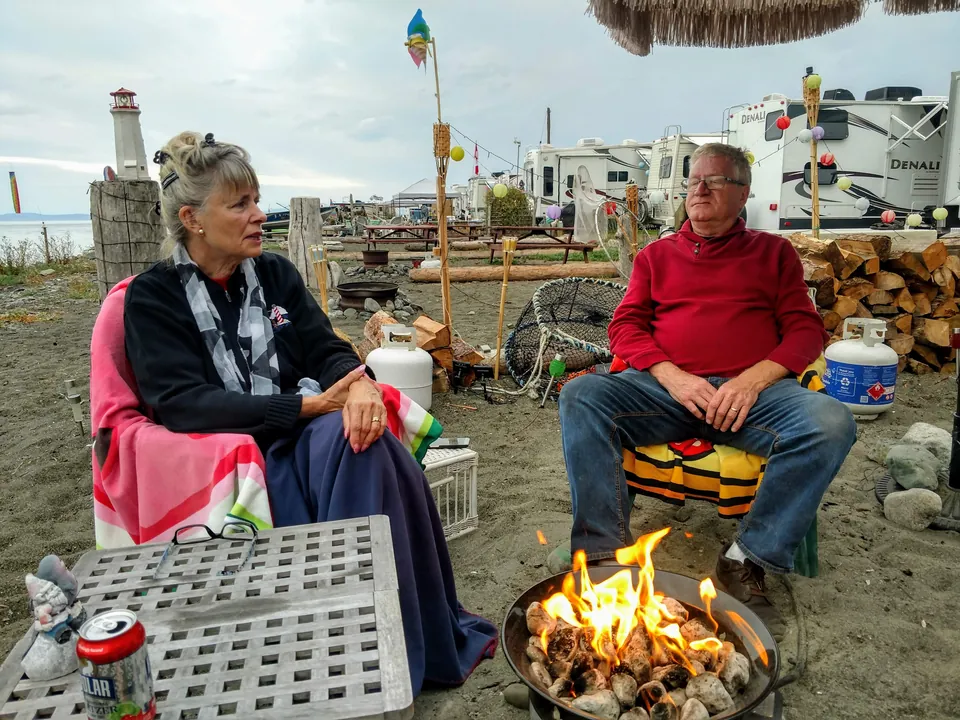 Twee gasten rond een kampvuur, met plaatsen aan het water op de achtergrond, bij Beachcomber RV Park