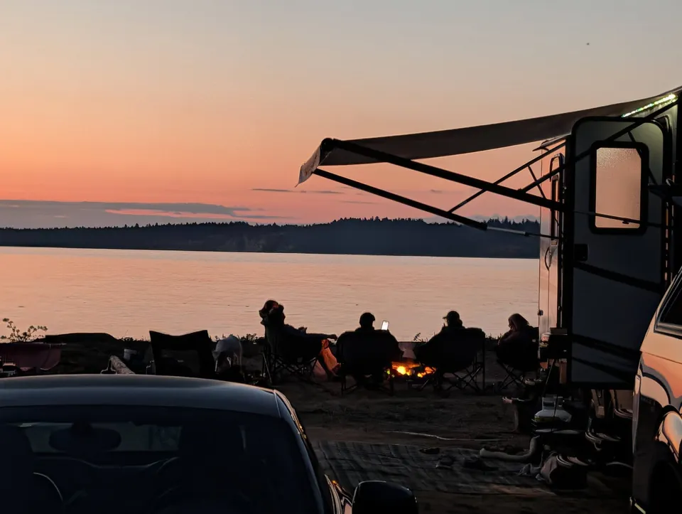 Gasten bij een camper en een klein vuur terwijl de lucht boven het water roze kleurt, bij Beachcomber RV Park