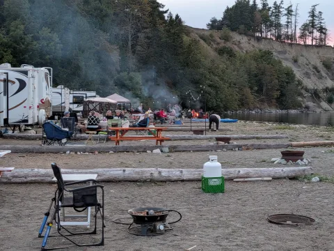 Campers relaxing beside RVs, picnic tables and fire rings below the shoreline bluffs at Beachcomber RV Park