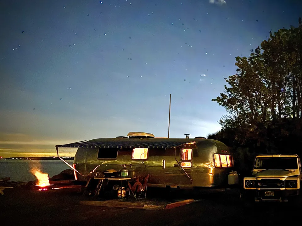 A silver travel trailer with glowing windows beside the water under a starry sky at Beachcomber RV Park